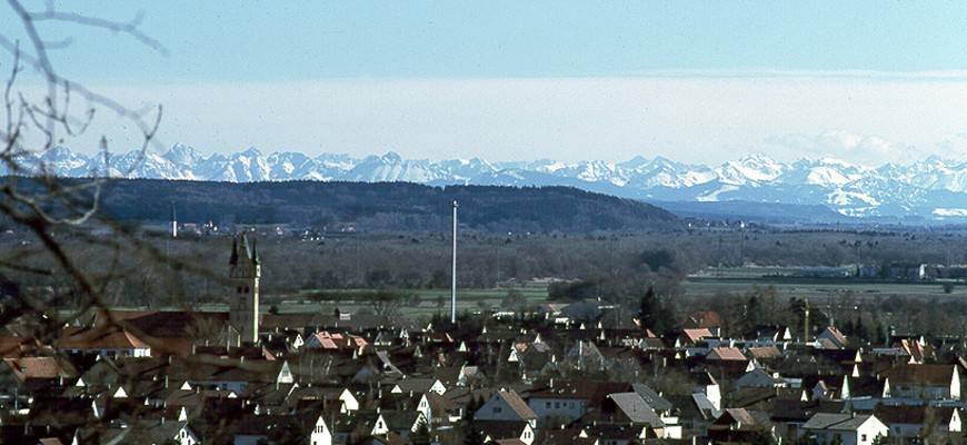 Häuser mit Kirchturm im Vordergrund vor einem Bergpanorama mit den entfernten Alpen im Hintergrund